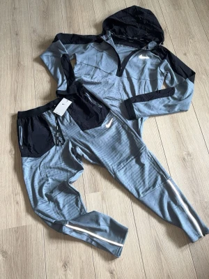 Nike phenom performance tracksuit (junior S/M) - Snyggt träningsset från Nike i grått med svarta detaljer. Setet består av en hoodie med dragkedja och matchande byxor med resårmidja och snörning. Båda plaggen har svarta fickor med dragkedja och reflekterande detaljer. Perfekt för träning eller chill. Storlek S/M för junior ungefär 160-170