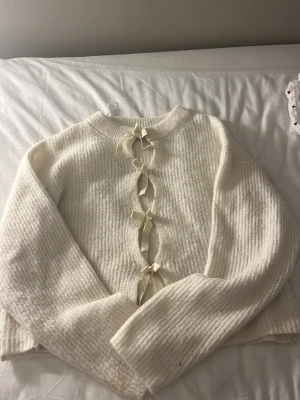 Vit stickad tröja med rosetter Gina Tricot - Supersöt vit stickad tröja från Gina Tricot med unika rosettdetaljer framtill. Tröjan har rund halsringning, långa ärmar och är gjord i ett mjukt, ribbstickat material. Perfekt för dig som vill ha något gulligt och trendigt i garderoben.