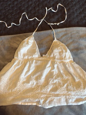 Vit broderad linnetopp med spets - Söt vit linnetopp med tunna axelband som knyts i nacken. Toppen har broderade detaljer och spetskant vid urringningen. Perfekt för varma dagar och ger en romantisk vibe till din outfit.
