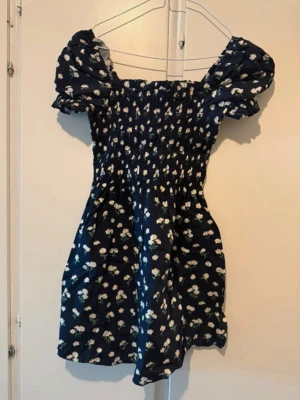Mörkblå blommig kortklänning - 🌸Supersöt mörkblå kortklänning med vita blommor och puffärmar. Klänningen har smock upptill och en figurnära passform i midjan. Perfekt för sommarens alla tillfällen och ger en romantisk vibe. Skick: använd en kväll