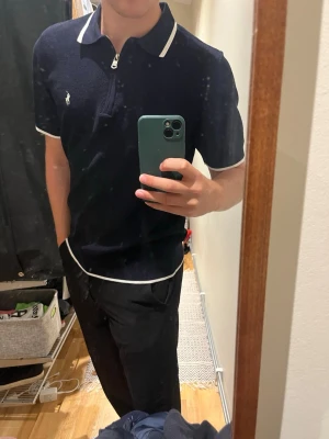 Marinblå pikétröja med vit kant - Snygg marinblå pikétröja med vit kant vid krage, ärmslut och nederkant. Tröjan har kort dragkedja framtill och en liten broderad logga på bröstet. Perfekt till jeans eller chinos för en clean look.