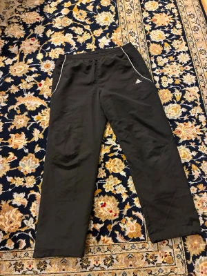 Svarta träningsbyxor från Adidas - Säljer ett par svarta träningsbyxor från Adidas med vita detaljer längs fickorna och Adidas-logga på vänster lår. Byxorna har rak passform och resår i midjan, perfekta för gymmet eller löprundan.