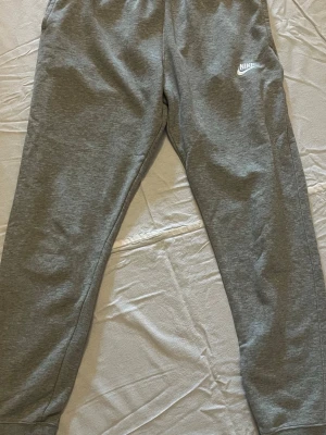 Gråa mjukisbyxor från Nike - Snygga gråa mjukisbyxor från Nike med klassisk vit logga på vänster ben. Byxorna har en avslappnad passform och är gjorda i mjukt bomullsmaterial. Perfekta för chill dagar eller när du vill ha det bekvämt.