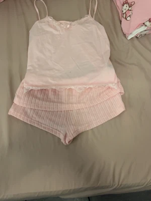 Ljusrosa pyjamas-set med spetsdetaljer - Sött ljusrosa pyjamas-set med linne och shorts. Linnet har tunna axelband och spetskant vid halsen samt nederkanten. Shortsen är randiga i samma ljusrosa ton och har en mjuk, bekväm passform. Perfekt för mysiga hemmakvällar.