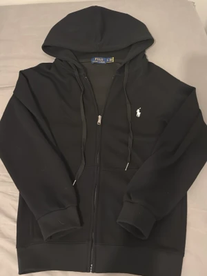 Polo zip Up hoodie - Snygg svart hoodie med dragkedja från Polo Ralph Lauren. Klassisk design med vit broderad logga på bröstet, huva med dragsko och två fickor framtill. Tillverkad i mjukt material som känns skönt mot huden. Perfekt för en avslappnad och stilren look. Den är i S men passar säkert i xs, den har aldrig kommit till användning eftersom den är för liten.