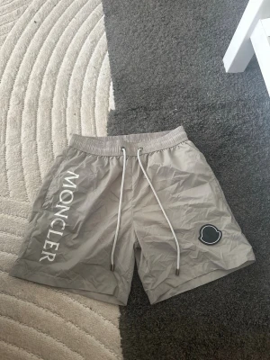 Gråa shorts från Moncler - Snygga gråa shorts från Moncler med vit logotyp längs ena benet och en patch på andra benet. De har elastisk midja med dragsko och är tillverkade i ett lätt, lite glansigt material. Perfekta för en avslappnad och trendig stil. funkar för S och M