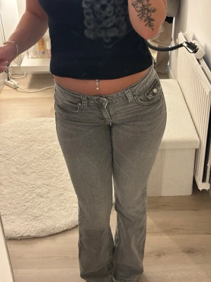 Grå lågmidjade bootcut jeans - Säljer ett par gråa lågmidjade jeans med bootcut passform. Jeansen har fram- och bakfickor med knappdetaljer och är tillverkade i ett mjukt bomullsmaterial. Perfekta för en avslappnad och trendig look.