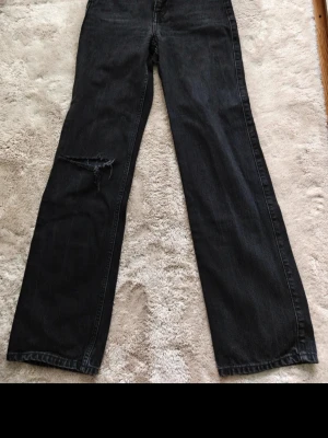 Svarta wide leg jeans från ROW - Säljer ett par svarta wide leg jeans från ROW med hög midja och en snygg sliten detalj på vänster ben. Jeansen har klassisk femficksdesign och dragkedja med knapp. Perfekta för en avslappnad och trendig look.