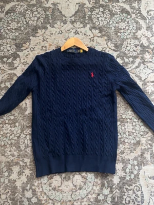 Mörkblå  tröja Polo Ralph Lauren - Kabelstickad mörkblå tröja från Polo Ralph Lauren med rund halsringning och den klassiska röda logotypen broderad på bröstet. Tröjan har långa ärmar och ribbade muddar vid ärmslut och nederkant. Perfekt för en stilren och tidlös look.