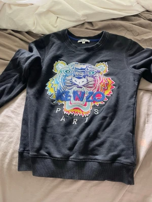 Svart sweatshirt med tiger från Kenzo - Cool svart sweatshirt från Kenzo med ikonisk broderad tiger i flera färger och texten 'Kenzo Paris' på bröstet. Tröjan har rund halsringning, långa ärmar och ribbade muddar.