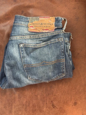 Blå jeans från Denim & Supply Ralph Lauren - Snygga blå jeans från Denim & Supply Ralph Lauren med klassisk femficksmodell och slitna detaljer. Jeansen har en rak passform och är gjorda i mjukt denimtyg med kontrastsömmar. Perfekta för en avslappnad och trendig look. I storlek W31 L32