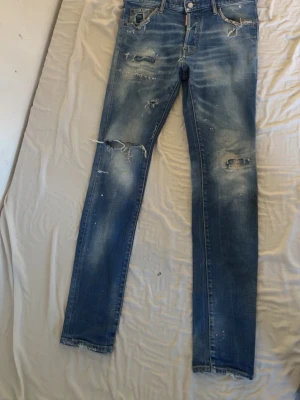 Blå slitna jeans från Dsquared2 - Säljer ett par blå jeans från Dsquared2 med distressed detaljer, slitningar och hål på benen. Jeansen har smal passform och klassisk femficksdesign. Materialet är denim i bomull och de har en cool, tvättad look med vita färgstänk.