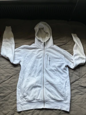 Vit hoodie från Sail Racing med dragkedja - Säljer en vit hoodie från Sail Racing med hel dragkedja framtill, huva och två sidofickor. Bröstficka med dragkedja och Sail Racing-logga. Insidan är mjuk och fleecefodrad för extra komfort. Perfekt för chill dagar eller när du vill ha en clean look.