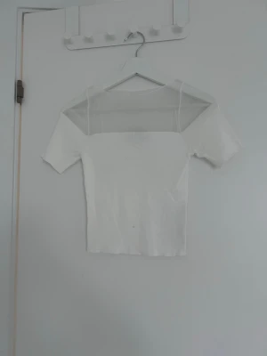 Vit topp med mesh-detalj - Säljer en vit topp med korta ärmar och transparent mesh-parti över axlar och bröst. Toppen har en rak, tight passform och är perfekt för dig som gillar stilrena plagg med en twist. Materialet känns mjukt och stretchigt.