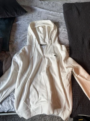  zip hoodie från Lacoste - Säljer en clean hoodie från Lacoste med dragkedja framtill och klassisk krokodil-logga på bröstet. Hoodien har fickor på sidorna, dragsko i huvan och är gjord i mjukt bomullsmaterial. Perfekt för en chill och stilren look.
