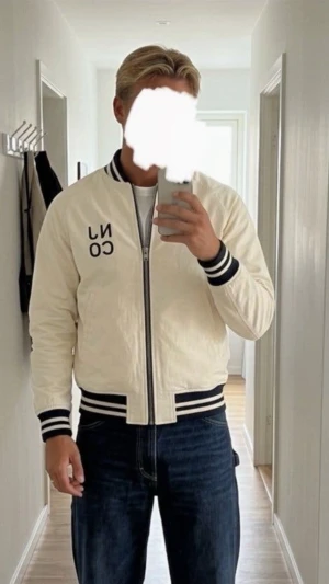 Vit och svart bomberjacka NJCO - Snygg bomberjacka i vitt och svart med broderad NJCO-text på bröstet. Jackan har randiga muddar vid ärmslut, krage och midja samt dragkedja framtill. Ärmarna är vita medan kroppen är svart, vilket ger en klassisk varsity-look. Perfekt för en trendig streetwear-stil.