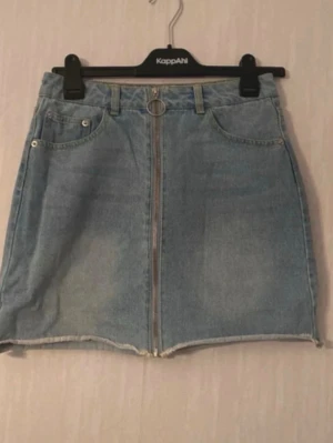 Blå jeanskjol med dragkedja - Snygg blå jeanskjol i kort modell med rå kant nertill och silverfärgad dragkedja framtill. Kjolen har fickor både fram och bak samt bälteshällor och en dekorativ ringdetalj. Perfekt för en trendig och avslappnad stil.