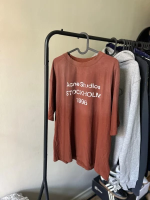 Brun Acne Studios t-shirt med tryck - Säljer en brun t-shirt från Acne Studios med vitt tryck framtill där det står 'Acne Studios STOCKHOLM 1996'. T-shirten har rund hals och trekvartsärm. Perfekt för dig som gillar stilrena plagg med en twist.