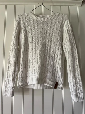 Vit kabelstickad tröja - Kabelstickad vit tröja med rund halsringning och långa ärmar. Tröjan har ribbade muddar vid ärmslut och nederkant samt ett klassiskt flätmönster över hela plagget. Perfekt för dig som gillar en stilren och mysig look.