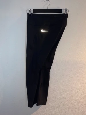 Svarta 3-kvarts träningstights från Nike - Snygga svarta 3-kvarts träningstights från Nike med hög midja och vit logga på låret. Tillverkade i stretchigt funktionsmaterial som passar perfekt för löpning, gym eller annan träning. Tightsen har en smickrande passform och diskreta sömmar. Finns ficka på låret som kan hålla telefon och en ficka baktill som kan hålla nycklar. 