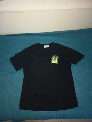 Svart Casablanca t-shirt med tryck - Säljer en svart t-shirt från Casablanca med färgglatt tryck på bröstet. T-shirten har klassisk passform och korta ärmar. Perfekt för dig som gillar stilrena plagg med en twist. Materialet är mjuk bomull.