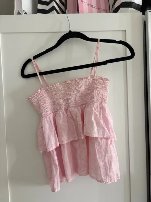 Ljusrosa volangtop från Gina Tricot - Supersöt ljusrosa topp från Gina Tricot med smock upptill och tunna axelband som man även kan ta bort. Den är använd 1-2 gånger! 