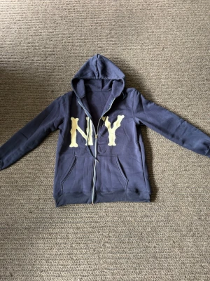 Marinblå hoodie med NY-tryck - Säljer en marinblå hoodie med dragkedja och stor huva. På framsidan finns ett stort gult NY-tryck i graffiti-stil. Hoodien har två fickor framtill och ribbade muddar vid ärmar och nederkant. Perfekt för en chill och avslappnad stil.