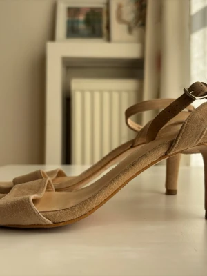 Beige sandfärgade sandaletter med smal klack från Rizzo storlek 36 - Snygga sandaletter i beige mocka med öppen tå och smal klack. Skorna har en enkel rem över tårna och justerbar ankelrem med spänne. Perfekt skick! Från Rizzo, storlek 36