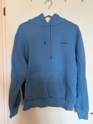Blå hoodie med tryck från AVNIIR S - Snygg blå hoodie från AVNIIR med minimalistisk logga på bröstet och stort grafiskt tryck på ryggen. Klassisk känguruficka framtill, ribbade muddar och justerbar huva med snören. Perfekt för dig som gillar streetwear och unika prints.