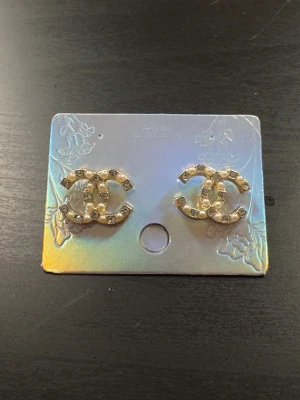 Guldfärgade örhängen med CC-logga, pärlor och strass  - Säljer ett par superfinna guldfärgade stud-örhängen formade som det klassiska CC-monogrammet. De är dekorerade med glittriga strasstenar och små vita pärlor.Skick: Helt nya / oanvända (sitter kvar på displaykortet). 