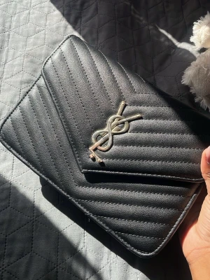 Svart YSL väska - Säljer min välanvända svarta YSL väska. Konstläder med silvrig kedjedetalj på framsidan. Väskan har ett klassiskt kuvertformat lock och diskreta sömmar i ett V-mönster. Perfekt att bära i handen eller över axeln för en stilren look. Ej äkta YSL där man kan se hur den guldiga färgen rostat lite, och hyffsat skadad innuti väskan. Den har svag kedja, där kedjan ibland lossnar men annars väldigt användbar.