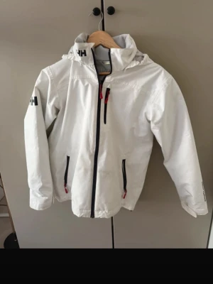 Vit vindjacka från Helly Hansen - Snygg vit vindjacka från Helly Hansen med huva och svarta dragkedjor. Jackan har tre fickor med dragkedja och logga på ärmen samt nacken. Perfekt för blåsiga dagar och enkel att matcha med olika outfits.