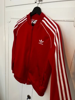 Röd Adidas track jacket med vita ränder - Klassisk röd Adidas track jacket med vita ränder längs ärmarna och vit Adidas-logga på bröstet. Dragkedja framtill, perfekt till en tracksuit look. Tillverkad i Primegreen-material för en sportig och retro look. Inga skador!