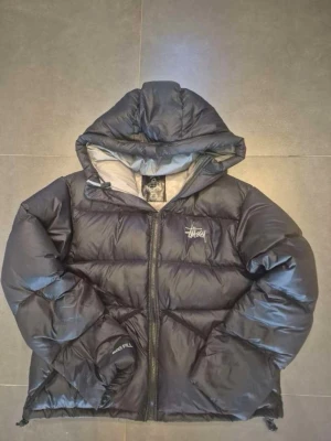 Stüssy 800 Fill Down Puffer Jacket - This is a black Stüssy puffer jacket with a hood. The front chest features the classic Stüssy logo in white embroidery. The sleeve has '800 FILL' embroidered, indicating the jacket uses 800 fill power down insulation, which is high quality and provides excellent warmth-to-weight ratio. The jacket has a full front zipper and appears to have a slightly shiny, water-resistant outer shell. The inside lining is a lighter color, possibly grey. This is a streetwear piece, popular for both style and functionality in cold weather.