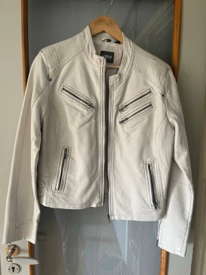 Vit skinnjacka från Livelo - Snygg vit bikerjacka från Livelo med flera dekorativa dragkedjor framtill och på bröstet. Jackan har en rak passform, ståkrage och detaljerade sömmar på axlar och ärmar. Materialet ser ut som konstläder och ger en cool look till din outfit. Storlek 46 är liten i storleken.