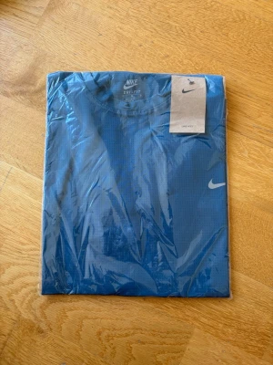 Blå Nike Dri-Fit t-shirt - Snygg blå Nike Dri-Fit t-shirt med klassisk rund hals och diskret Nike-logga på bröstet. Tillverkad i ett lätt och funktionellt material som andas, perfekt för träning eller en aktiv vardag. Enkel och stilren design med korta ärmar.