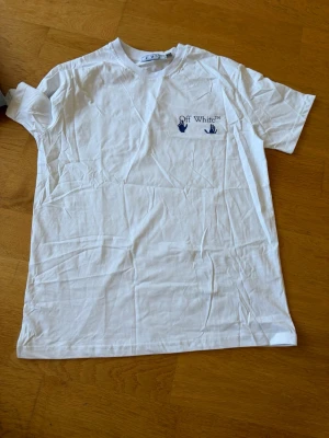 Vit Off-White t-shirt i bomull - Säljer en vit t-shirt från Off-White med blå logga och handtryck på bröstet. Klassisk rund hals och korta ärmar. Tillverkad i mjuk bomull som känns skön mot huden. Perfekt till jeans eller shorts för en clean och enkel look.