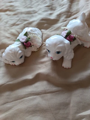 Söta Vintage Hundar i Porslin – Romantisk Kitsch & Coquette! - Säljes: Två otroligt charmiga porslinsfigurer i form av söta hundar/pudlar! De är söta dekorativa detaljer med fina handmålade ansiktsdrag, söta tygblommor och pärlhalsband runt halsen. Perfekta för dig som älskar vintage, kitsch eller coquette-estetik. Innehåll: 2 st porslinsfigurer (säljs som ett par). Märkning: Made in Taiwan (klistermärke finns kvar under) Skick: I mycket fint skick utan nagg eller sprickor (Se bilder för egen bedömning) Skicka meddelande vid frågor. 