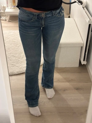 Blå bootcut jeans med låg midja - Säljer ett par klassiska blå bootcut jeans med låg midja och fem fickor. Jeansen har en snygg tvätt och är tillverkade i stretchigt denimtyg som sitter bekvämt. Perfekta för dig som gillar en avslappnad men ändå trendig look.