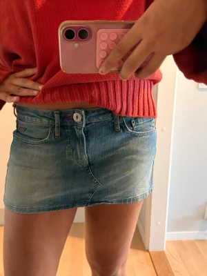 Blå jeanskjol med fickor - Säljer en kort blå jeanskjol med klassisk femficksdesign och knappstängning fram. Kjolen har en patch med text på ena bakfickan och bälteshällor i midjan. Perfekt för dig som gillar en avslappnad och trendig stil.