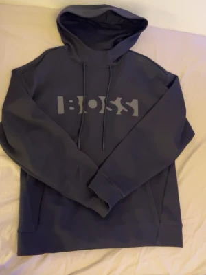 Mörkblå hoodie från BOSS - Snygg mörkblå hoodie från BOSS med stor logga framtill. Klassisk modell med huva och dragsnören samt känguruficka. Tillverkad i mjukt material som känns skönt mot huden. Perfekt för en avslappnad och trendig look.