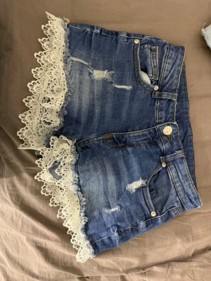 Blå jeansshorts med spetsdetalj - Säljer ett par blå jeansshorts med slitningar och snygga vita spetsdetaljer längs bensluten. Shortsen har klassisk femficksmodell, knapp och dragkedja framtill samt en midwaist passform. Perfekta för sommaren och ger en cool men söt vibe.