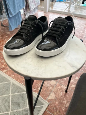 Svarta lanvin skor med lackdetalj - Säljer ett par svarta lanvin med snygg lackad tå och mockaliknande material på sidorna. Skorna har vita sulor och svarta skosnören. Perfekta för dig som vill ha en stilren men ändå unik look.