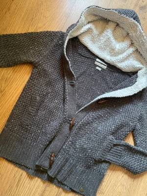 Grå stickad kofta med huva - Mysig grå stickad kofta från Lee Cooper med luva fodrad i mjuk teddy. Koftan har träknappar, två fickor framtill och ribbade muddar. Perfekt för kyliga dagar och ger en avslappnad look.
