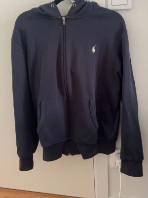Mörkblå polo zip hoodie - Snygg mörkblå hoodie från Polo Ralph Lauren med dragkedja framtill och klassisk vit logga på bröstet. Hoodien har huva med snörning, två fickor fram och ribbade muddar vid ärmar och nederkant. Tillverkad i mjukt bomullsmaterial för skön känsla.