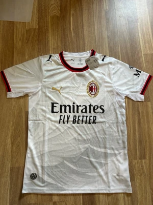 Vit AC Milan matchtröja  2026/27 - Säljer en vit AC Milan matchtröja från Puma med klubbmärke på bröstet och svart Emirates Fly Better-tryck. Tröjan har röda och svarta detaljer vid krage och ärmslut samt korta ärmar. Tillverkad i ett lätt funktionsmaterial, perfekt för fotbollsträning.