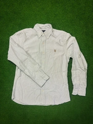 Randig Ralph lauren skjorta  - Klassisk vit skjorta med tunna blåa ränder och knappar framtill. Skjortan har lång ärm, krage och en liten broderad logga på bröstet. Perfekt för en clean och stilren look.