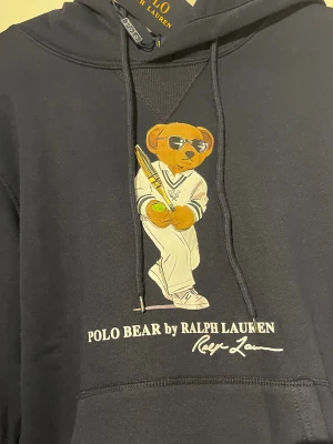Mörkblå hoodie Polo Bear Ralph Lauren - Mörkblå hoodie från Ralph Lauren med Polo Bear-tryck framtill där björnen har solglasögon och håller ett tennisracket. Klassisk känguruficka, dragsko i huvan och ribbade muddar. Snygg och sportig design som sticker ut.
