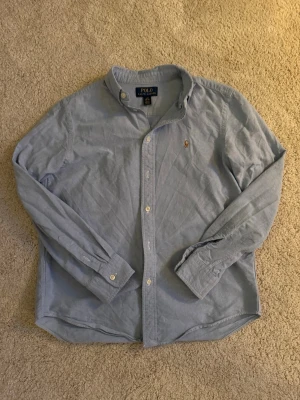 Ljusblå skjorta från Polo Ralph Lauren - Klassisk ljusblå skjorta från Polo Ralph Lauren med button-down krage och broderad logga på bröstet. Skjortan har långa ärmar, vita knappar och är i väldigt bra skick. Perfekt för en clean och stilren look. Det är en skjorta i barn storlek 14-16 (160). 