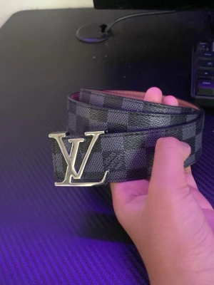 Bälte - Snyggt bälte från Louis Vuitton med klassiskt rutmönster i svart och grått. Bältet har en stor LV-logga i silver som spänne och är gjort i ett tåligt material med exklusiv känsla. Perfekt accessoar för att lyfta din outfit.